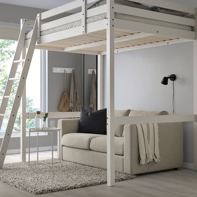 A white STORÅ loft bed with a ladder on the left, larger than surrounding furniture, featuring a sofa below.