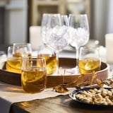 Four wine glasses of different styles on a wooden tray with pistachios and dried leaves. Glasses and tray on a wooden table with a cream cloth.