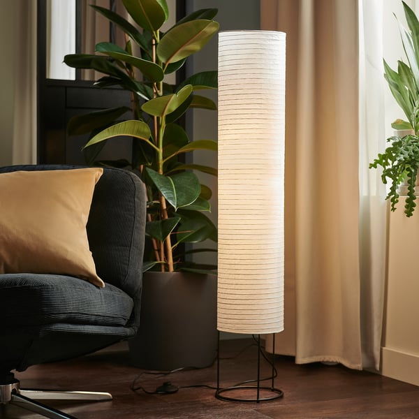 Floor lamp with white shade, slim black base, beside chair and plant.