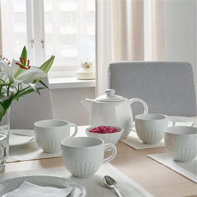 Table set with white STRIMMIG mugs, teapot, plates, and cutlery. Flowers in a vase beside the teapot. Soft lighting creates a cozy atmosphere.