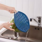 Person pours water from a strainer bowl into a stainless steel sink. The strainer bowl is blue with a handle.