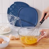A person is using a glass bowl with a lid and whisk to mix egg yolks. The bowl set is versatile for preparing and storing food.