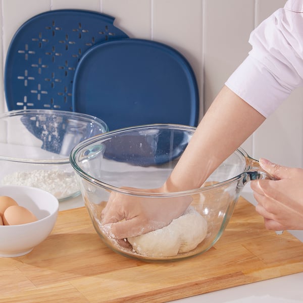Person mixing dough in glass bowl on wooden board, with clear and blue covers nearby.