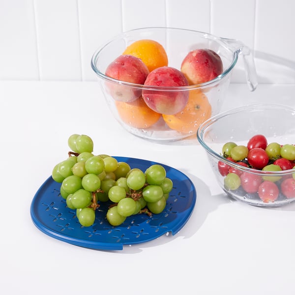 Three glass bowls hold fruit; oranges, apples, grapes, and tomatillos. One sits on a blue strainer, demonstrating food preparation.