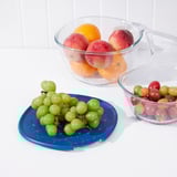 Three glass bowls hold fruit; oranges, apples, grapes, and tomatillos. One sits on a blue strainer, demonstrating food preparation.
