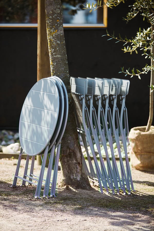 Stack of blue folding chairs, placed outdoors against a dark wooden fence, beneath a tree.