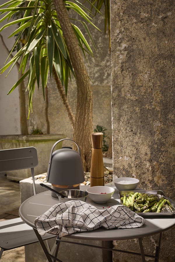Outdoor setup with SUNDSÖ table and chair, featuring kitchen tools, bowls, and greens on a grey table with a palm tree and stone wall backdrop.