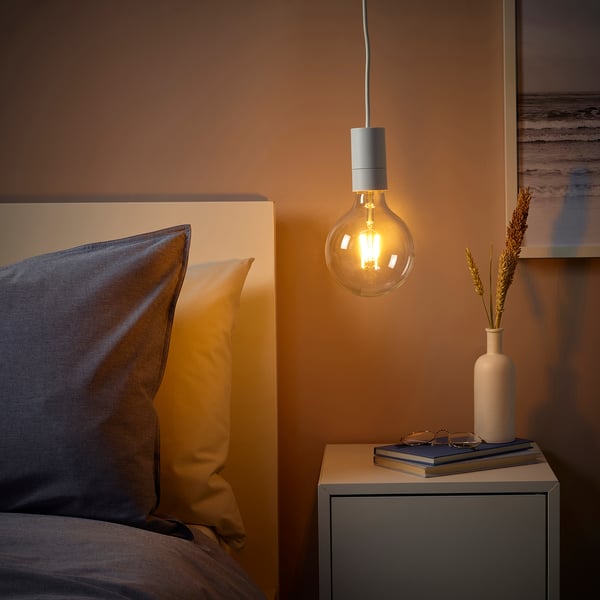 Cozy bedroom corner with SUNNEBY lamp, orange bulb, blue & yellow pillow, bedside table with book & vase, warm brown wall & modern painting.