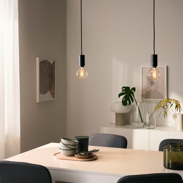 Two SUNNEBY pendant lamps over dining table with grey chairs, bowls, and kitchen roll. Abstract art, white lamp, and plants on wall.