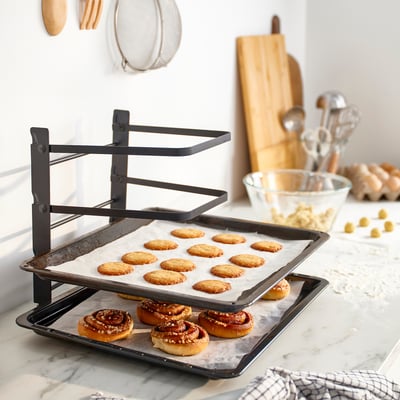 Black SVARTABBORRE baking tray rack holds 2 trays, shown with cookies. Folds flat for easy storage.