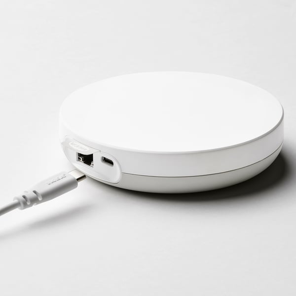 A white wireless charging pad with usb ports connects to ikea home smart app for controlling speakers, playing/pausing, and adjusting volume.