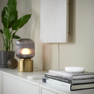 A modern display a small white SYMFONISK sound remote on a stack of books, next to a stylish lamp and a plant.