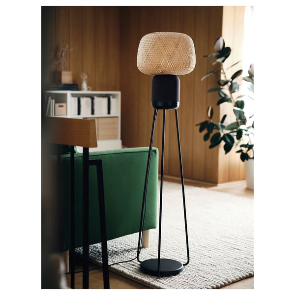 Black SYMFONISK floor lamp speaker on a woven rug near a green chair. Modern design with bamboo shade, emitting warm light.
