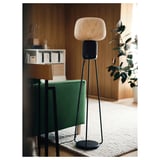 Black SYMFONISK floor lamp speaker on a woven rug near a green chair. Modern design with bamboo shade, emitting warm light.