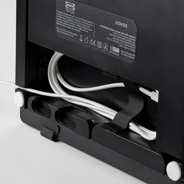 A black ikea SYMFONISK wifi speaker with its back panel open, revealing internal cables and a label with product details.