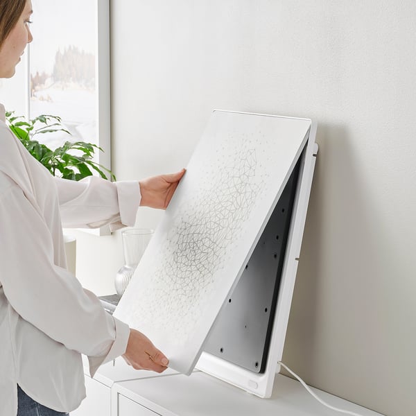 A person opens a white picture frame speaker, revealing its modern design and positioning it on a white stand next to a plant.