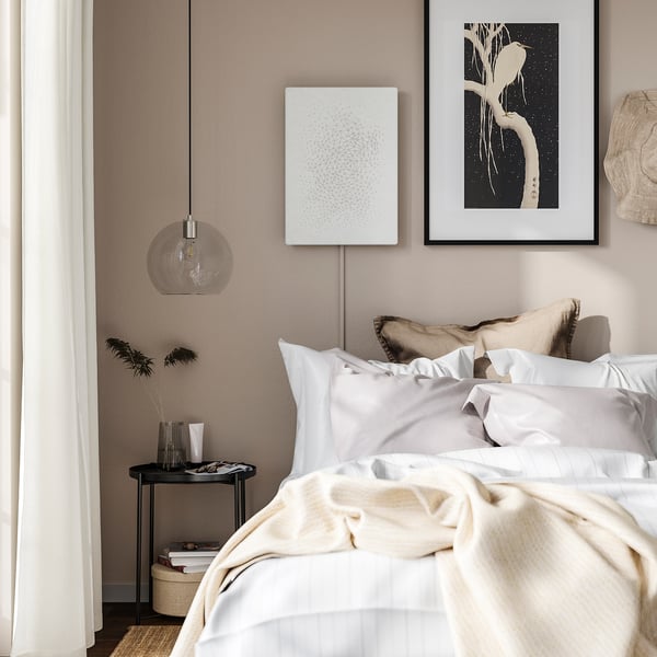 Elegant bedroom: white bed, cream walls, hanging lamp, art, side table with books, vase.