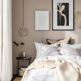 Elegant bedroom: white bed, cream walls, hanging lamp, art, side table with books, vase.