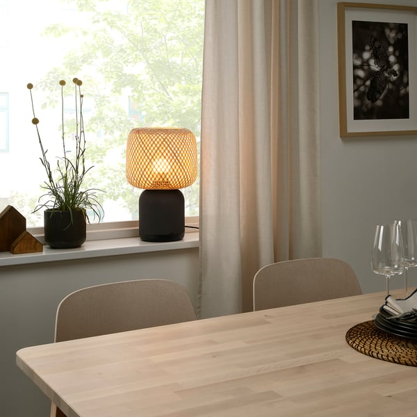 Small bamboo shade on speaker lamp sits near window, between curtains and chairs, beside a potted plant.