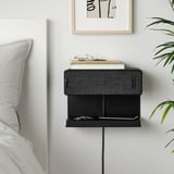 Small black SYMFONISK wall-mounted speaker & shelf combo next to bed. Houses books, holds phone for charging. Modern, minimal design.