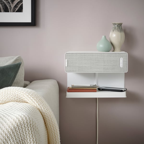 SYMFONISK white bookshelf speaker with wireless charger, on wall. Near cozy sofa with blanket, books, and decor.