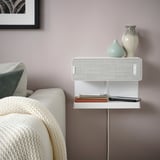 SYMFONISK white bookshelf speaker with wireless charger, on wall. Near cozy sofa with blanket, books, and decor.