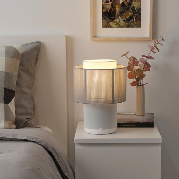 A cozy bedroom with SYMFONISK wifi speaker lamp, gray vase of dried flowers, book, and patterned pillows.