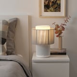 A cozy bedroom with SYMFONISK wifi speaker lamp, gray vase of dried flowers, book, and patterned pillows.
