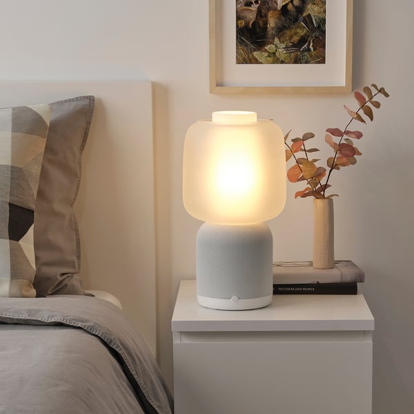 Modern bedroom with SYMFONISK lamp-speaker, vase of autumn leaves, book, cozy bed, patterned cushions, grey blanket.