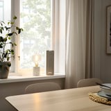 Modern dining area with wooden table, white chairs, white lamp, tall beige SYMFONISK speaker, green plant, window with curtains, and table setting.
