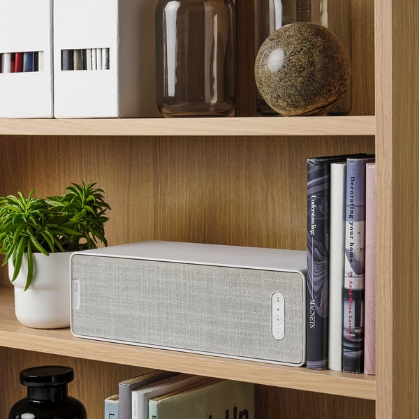 A white SYMFONISK speaker sits on a wooden shelf, flanked by books and a plant. It streams music via wifi and integrates with other sonos products.
