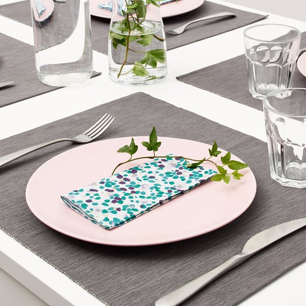 Table setting with pink plates, colorful pencils napkins, silverware, and glass cups. Greenery in vases adds decor.