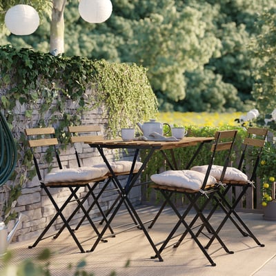 Small patio set with wooden table, 4 chairs, cushions. Surrounded by greenery, cozy outdoor seating.