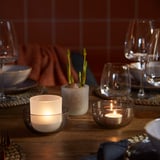Wooden table set with lit tealights in glass holders, wine glasses, plates, and small potted plant.