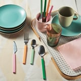 Colorful cutlery set on a perforated tray, with plates, cups, and glasses. Forks, knives, spoons, and teaspoons are neatly arranged, ready for use.