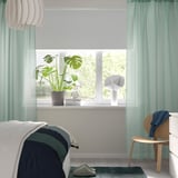 Green curtains frame a window with plant, books, lamp. Pool above floor, natural light. Minimalist room with bed, chair, slippers.