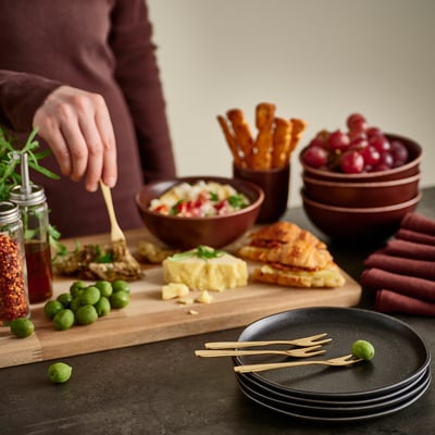 A person seasoning food on a wooden board with TILLAGD forks and bowls of snacks nearby.