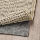 A beige woven rug with dark accents, placed on a gray non-slip pad on a wooden floor.