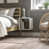 Cozy bedroom with wooden bed, white bedding, floating shelf, wicker chair, beige rug.