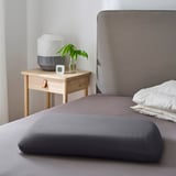 Bedroom scene with ergonomic pillow on bed, beside table with lamp, plant, and clock.
