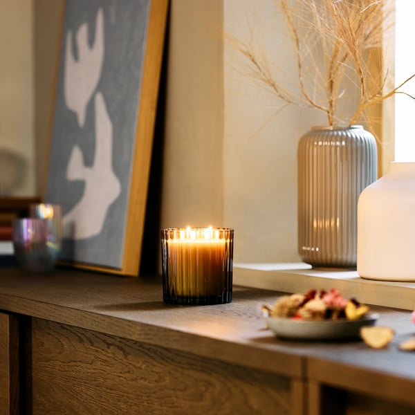 A cosy living space with TOFSTÄRNA scented candle. Lit candle on shelf with decor, vases, and abstract art.