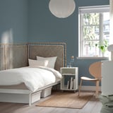 Bedroom with TOLKNING blue rattan headboard, white bedding, wooden chair, side table with books and window.
