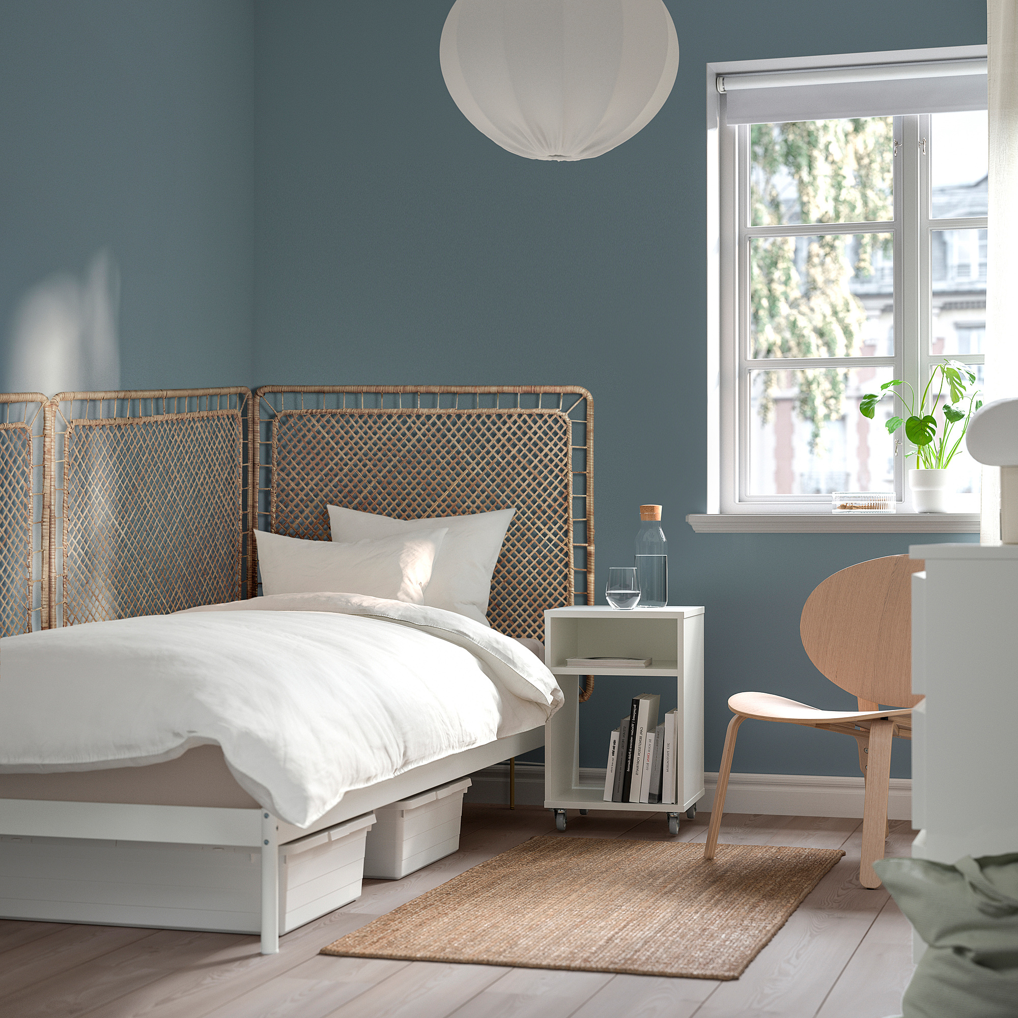 Bedroom with TOLKNING blue rattan headboard, white bedding, wooden chair, side table with books and window.