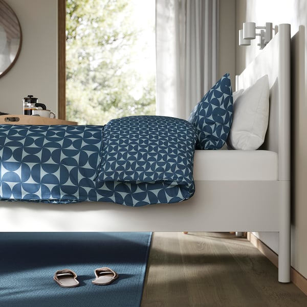 A modern bedroom with a TONSTAD bed featuring a white frame, blue patterned bedding, and slippers placed on a blue rug beside it.