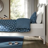 A modern bedroom with a TONSTAD bed featuring a white frame, blue patterned bedding, and slippers placed on a blue rug beside it.