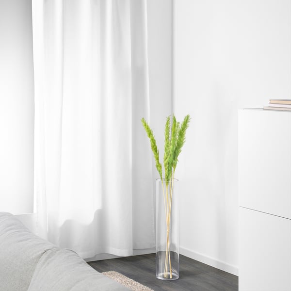 A minimalist room featuring a tall, clear vase with dried plants, positioned near a white curtain and a gray sofa, adding a touch of natural decor.