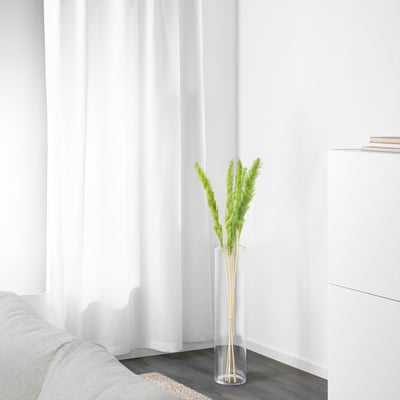 A minimalist room featuring a tall, clear vase with dried plants, positioned near a white curtain and a gray sofa, adding a touch of natural decor.