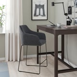 Modern gray TOSSBERG bar stool with padded seat and back, beside a desk with laptop and plant.