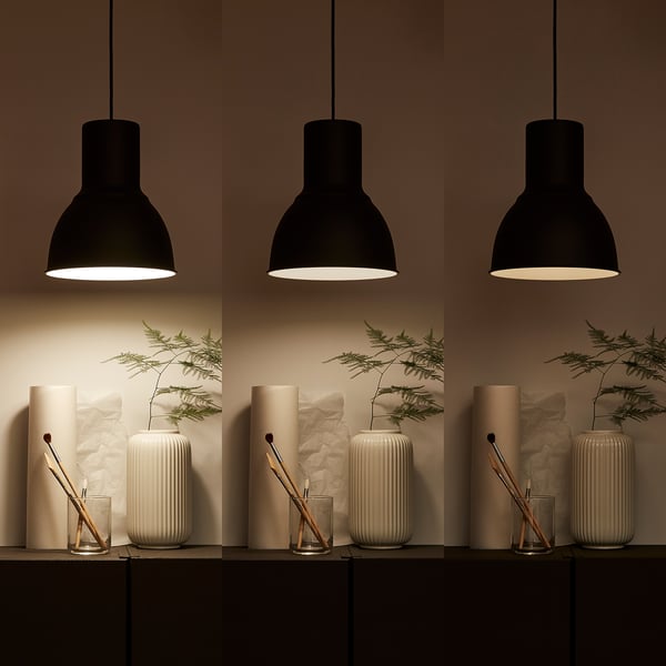 Three hanging black lamps illuminate a shelf with cylindrical white vases holding green branches and small glass containers with brushes.