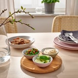 A dining table set with TRÅDTICKA bowls, containing snacks and sauces, placed on a wooden tray. Accompanied by stacked plates and cutlery.
